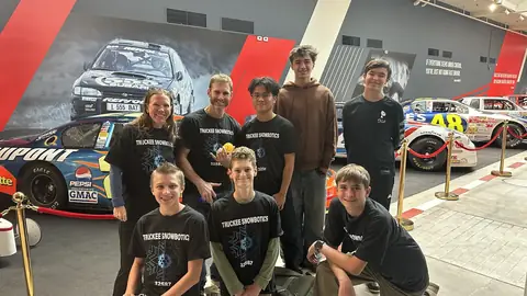 Team members and mentors in Truckee Snowbotics shirts in front of a display of race cars.